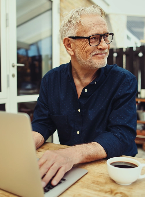 A man wearing glasses while working on a laptop with a cup of coffee.