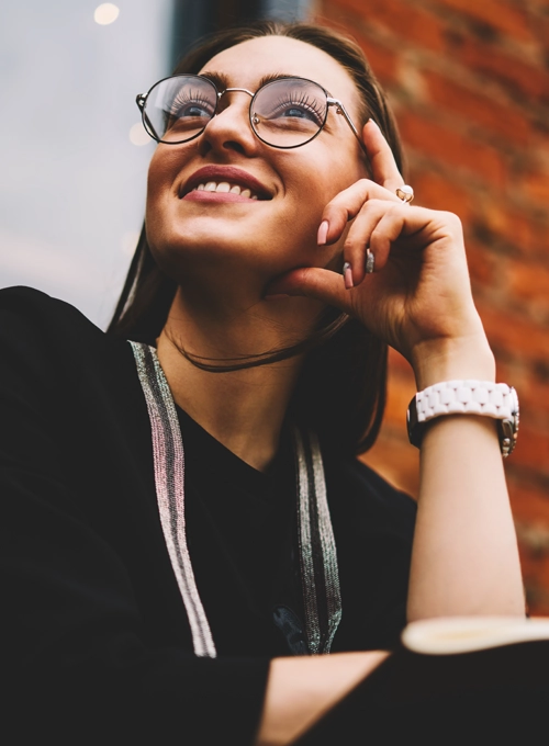 A woman smiling with glasses on while sitting in front of a brick building.