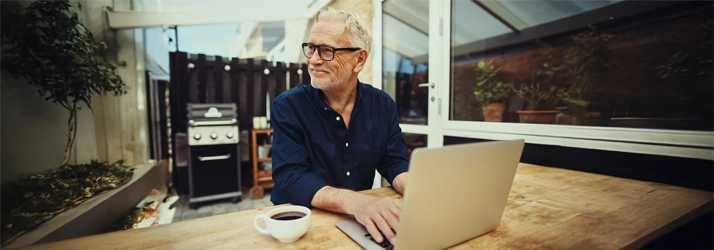 A man smiling while working on a laptop while wearing glasses.